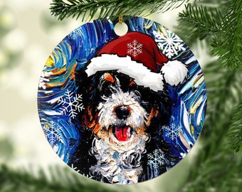 May include: Round Christmas ornament featuring a Bernese Mountain Dog wearing a Santa hat. The dog's portrait is set against a blue and white textured background with snowflake accents. A festive holiday decoration.