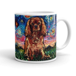 May include: A white ceramic coffee mug with a colorful painting of a Cavalier King Charles Spaniel dog. The dog is sitting in a field of grass with a starry night sky in the background. The painting is done in a style reminiscent of Vincent van Gogh.