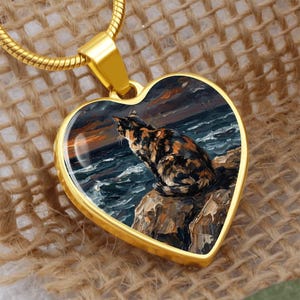 May include: Gold-toned heart-shaped pendant featuring a painting of a cat perched on a rock overlooking a stormy sea. The cat has a tortoiseshell pattern. The pendant hangs from a gold chain.