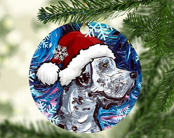 May include: Round Christmas ornament featuring a dog wearing a Santa hat. The dog is painted in shades of gray and white, set against a background of blue and pink brushstrokes. White snowflakes are scattered around the dog's head.