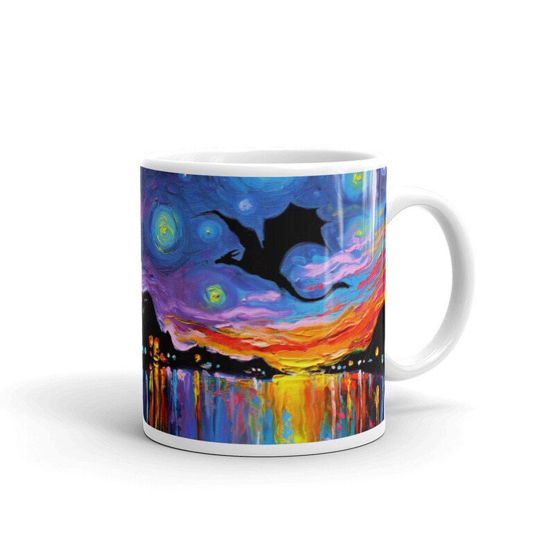 May include: A white ceramic mug with a colorful painting of a silhouette of a dragon flying over a city skyline at sunset. The sky is a vibrant blue with yellow, orange, and purple clouds. The city lights are reflected in the water below.