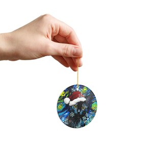 Pu&ograve; includere: Un ornamento rotondo in ceramica raffigurante un gatto nero con un cappello da Babbo Natale rosso e bianco. Lo sfondo presenta un motivo a notte stellata con vortici blu, gialli e verdi e fiocchi di neve bianchi. Appeso a un filo dorato.