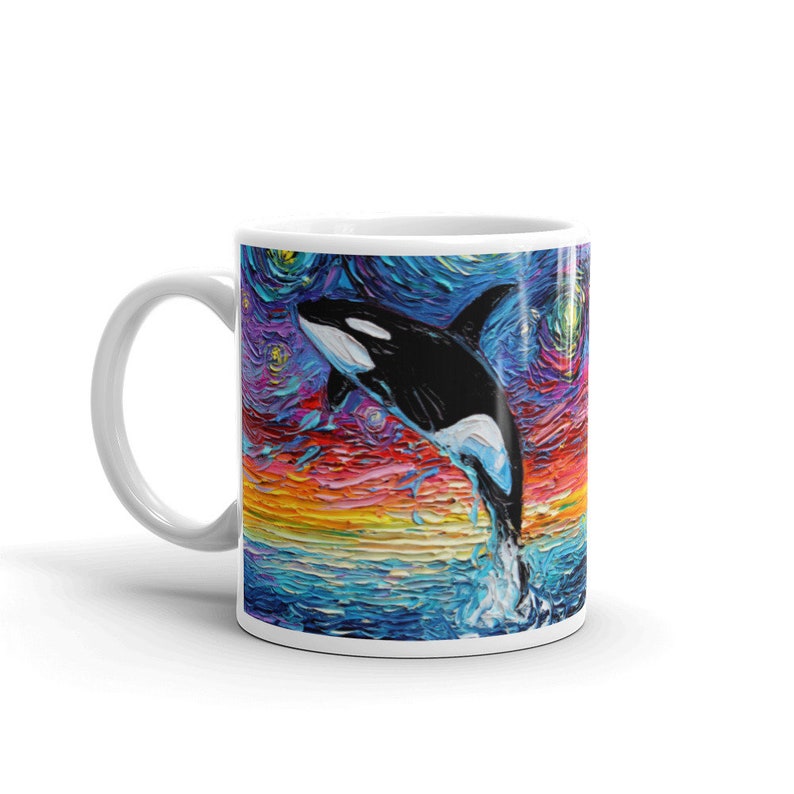 K&ouml;nnte beinhalten: Wei&szlig;e Keramiktasse mit einem farbenfrohen Gem&auml;lde eines Orcas, der aus dem Wasser springt. Der Hintergrund ist ein sternen&uuml;bers&auml;ter Nachthimmel mit Sonnenuntergang.