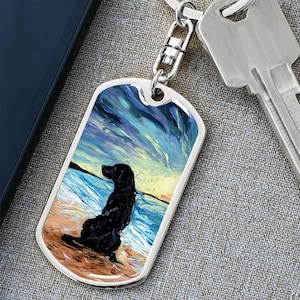 May include: A silver dog tag keychain featuring a black dog sitting on a beach with a colorful sky and ocean background. The dog tag is attached to a key and a silver chain.