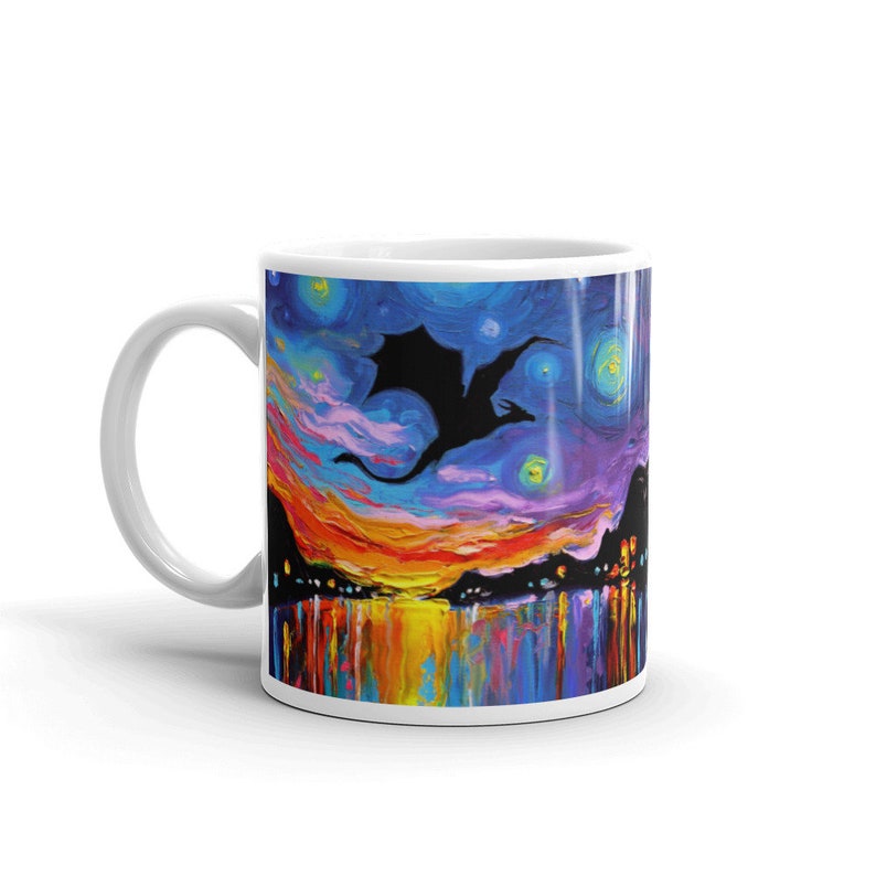May include: White ceramic mug with a colorful painting of a black silhouette of a dragon flying in a starry night sky with a reflection in the water below.