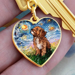 May include: A gold-colored heart-shaped pendant featuring a painting of a brown dog in a field under a starry night sky. The pendant is attached to a gold-colored key.
