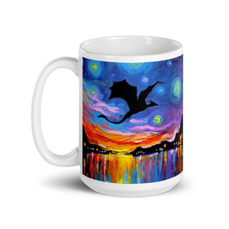 May include: White ceramic mug with a colorful painting of a black silhouette of a dragon flying through a starry night sky. The sky is painted in shades of blue, purple, and orange, with a reflection of the sky in the water below.