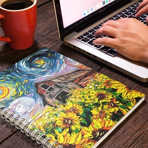 May include: A spiral-bound notebook with a colorful painting of sunflowers and a starry night scene. A red coffee mug and a laptop with hands typing on the keyboard are also visible on a wooden surface.