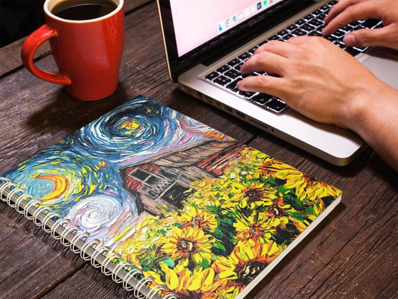 May include: A spiral-bound notebook with a colorful painting of sunflowers and a starry night scene. A red coffee mug and a laptop with hands typing on the keyboard are also visible on a wooden surface.