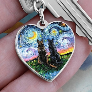 May include: Heart-shaped pendant with a colorful painting of two dogs sitting on a grassy hill under a starry night sky. The pendant is attached to a silver key.