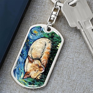 May include: A silver-colored dog tag keychain featuring a sleeping cat in a painting style reminiscent of Van Gogh's "Starry Night." The cat is orange and white, resting on green grass with a blue sky background. A key and a phone are in the background.