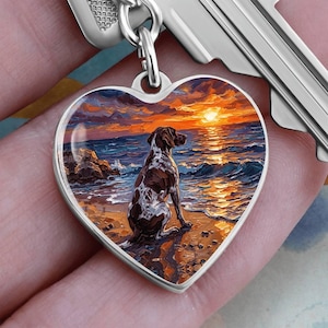 Puede incluir: Colgante en forma de corazón con una ilustración de perro. El colgante presenta un perro marrón y blanco sentado en una playa, mirando una puesta de sol sobre el océano. El colgante está unido a una llave plateada.