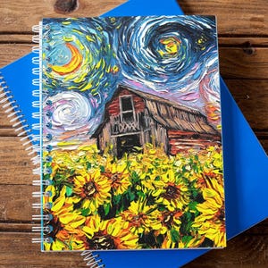May include: A spiral-bound notebook featuring a vibrant painting of a barn in a field of sunflowers under a starry night sky. The notebook is set against a blue notebook on a wooden surface.