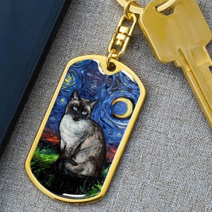 Può includere: Targhetta dorata per animali domestici con un'illustrazione di un gatto siamese su uno sfondo di cielo stellato. Il gatto ha gli occhi blu ed è seduto sull'erba verde. Una chiave d'oro e un telefono sono sullo sfondo.