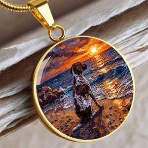 May include: Gold-toned pendant necklace featuring a round charm with a painted scene of a dog on a beach at sunset. The dog is brown and white, facing the ocean. The background shows a vibrant sunset with orange, yellow, and blue hues.
