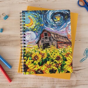 May include: Spiral-bound notebook featuring a colorful painting of a barn in a field of sunflowers under a swirling night sky. The notebook is on top of a yellow notepad, with blue and red pens, blue paper clips, and scissors nearby.
