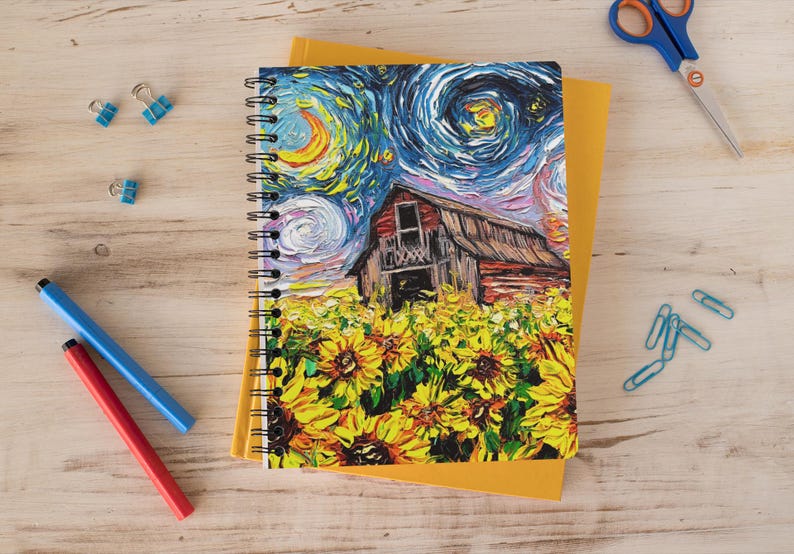 May include: Spiral-bound notebook featuring a colorful painting of a barn in a field of sunflowers under a swirling night sky. The notebook is on top of a yellow notepad, with blue and red pens, blue paper clips, and scissors nearby.