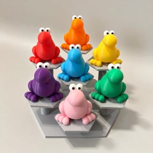 May include: A display of seven colorful frog figurines in red, orange, yellow, blue, green, purple, and pink. Each frog has large white eyes and is posed on a gray hexagonal shelf. The frogs are a whimsical decorative item.