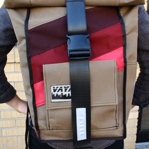 May include: A tan, red and pink canvas backpack with a black adjustable strap and a white reflective strip. The backpack has a large pocket on the front with a black and white logo that says "KAYZ".