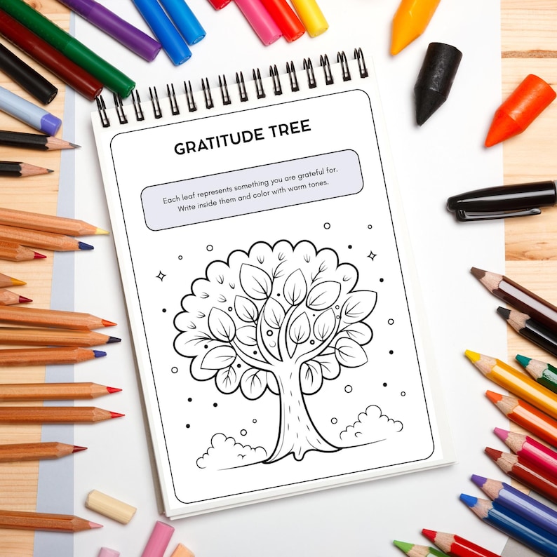 May include: A spiral-bound notebook with the title "GRATITUDE TREE" and a tree illustration. The text encourages users to write and color the leaves. Colored pencils and crayons surround the notebook, suggesting a creative activity.