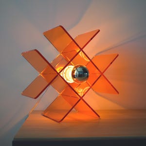 Può includere: Una lampada da tavolo geometrica arancione con una lampadina argentata. La lampada ha un design unico con pannelli angolati e intersecanti. La lampada è accesa, proiettando un bagliore caldo. La lampada è su una superficie di legno.