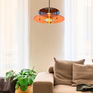 Może przedstawiać: Nowoczesna lampa wisząca o wielu warstwach. Lampa posiada okrągły, pomarańczowy klosz z mniejszym, ciemnym dyskiem nad nim. W środku wisi kulista, podświetlona żar&oacute;wka, emitująca ciepłą poświatę. Światło wisi na czarnym kablu.