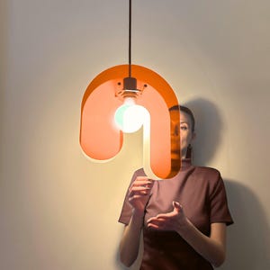 Könnte beinhalten: Orange-weiße Pendelleuchte mit geschwungenem Design. Die Leuchte hat eine große, abgerundete Form mit einer Glühbirne in der Mitte. Die Leuchte hängt an einem schwarzen Kabel.