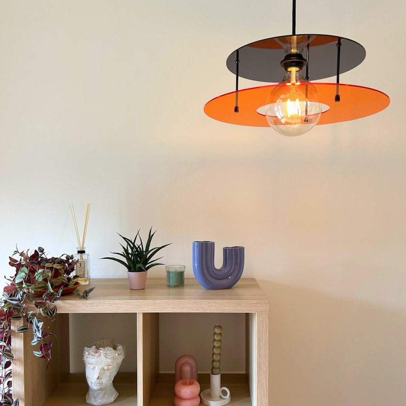 Może przedstawiać: Nowoczesna lampa wisząca z dwoma poziomami, z czarnym g&oacute;rnym dyskiem i żywym pomarańczowym dolnym dyskiem. Lampa wisi nad drewnianą p&oacute;łką z elementami dekoracyjnymi, w tym niebieskim wazonem i roślinami doniczkowymi. Żar&oacute;wka jest widoczna.