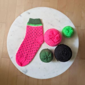 May include: A knitted sock with a watermelon design, featuring a pink body with black seed patterns and a green cuff with a lime stripe. Several balls of yarn in pink, green, and black are arranged nearby, suggesting a knitting project.