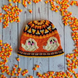 May include: A knitted orange, black, and white hat with a candy corn pattern and two white ghosts. The hat is surrounded by candy corn.