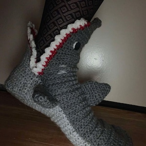 Puede incluir: Zapatillas de tiburón tejidas a mano en tonos grises, con un ribete blanco y rojo alrededor de la boca abierta. Las zapatillas presentan una aleta en el lateral y un ojo negro, creando un diseño lúdico. Las zapatillas están sobre un suelo de madera.