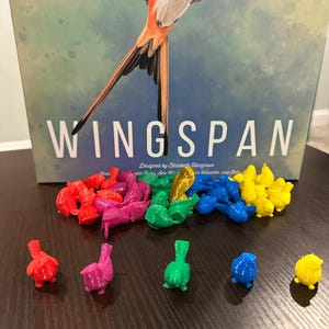 May include: A collection of colorful bird figurines, including red, pink, blue, green, and yellow. The figurines are arranged on a wooden surface. The game "Wingspan" is visible in the background.