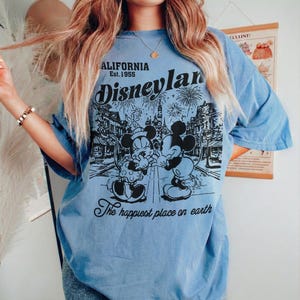 May include: Light blue t-shirt featuring a black and white graphic of Mickey and Minnie Mouse in front of a cityscape with fireworks. The shirt has the text "CALIFORNIA Est. 1955 Disneyland" and "The happiest place on earth."