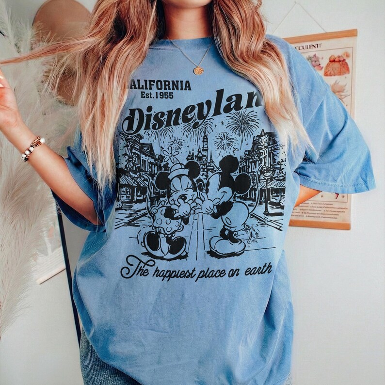 May include: Light blue t-shirt featuring a black and white graphic of Mickey and Minnie Mouse in front of a cityscape with fireworks. The shirt has the text "CALIFORNIA Est. 1955 Disneyland" and "The happiest place on earth."