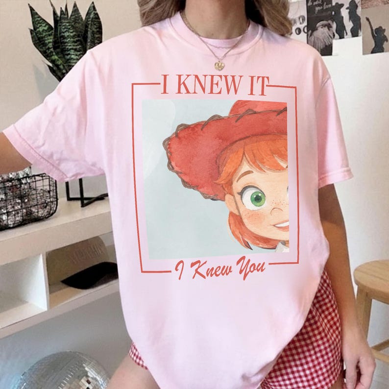 以下が含まれることがあります： 上部に「I KNEW IT」、下部に「I Knew You」というテキストが書かれた長方形のグラフィックがプリントされたライトピンクのTシャツ。グラフィックには、赤い髪、赤い帽子、緑色の目をしたキャラクターの水彩画風のイラストが描かれています。