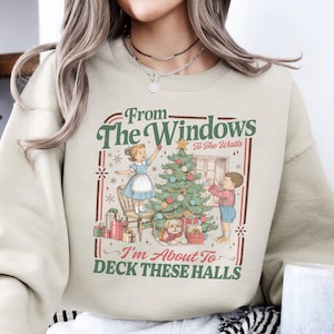 Puede incluir: Una sudadera beige con un gráfico navideño vintage. El gráfico presenta a niños decorando un árbol de Navidad, con el texto "From The Windows To The Walls I'm About To Deck These Halls."