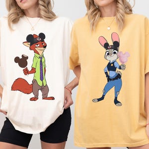 Puede incluir: Dos camisetas con personajes de dibujos animados. Una es de color crema con un zorro con un sombrero de Mickey Mouse y sosteniendo un helado con forma de Mickey Mouse. La otra es amarilla con un conejo con uniforme de polic&iacute;a sosteniendo algod&oacute;n de az&uacute;car.