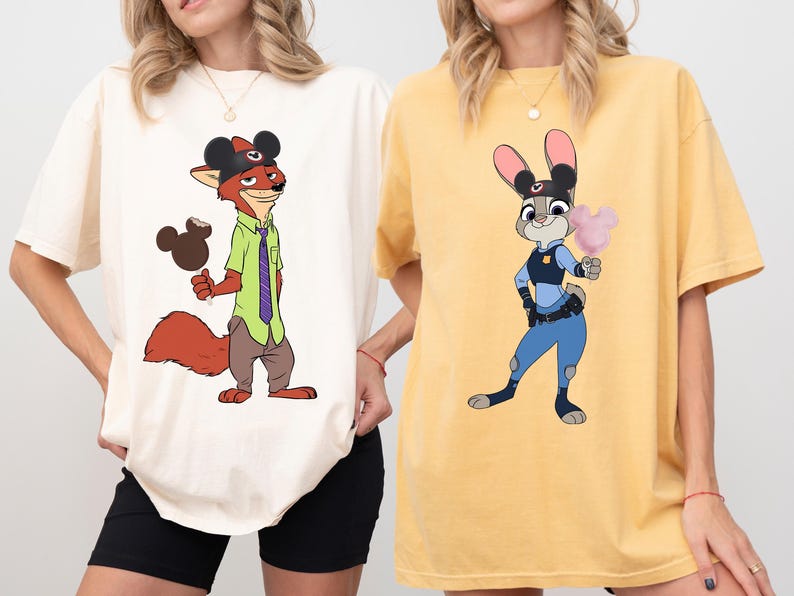 Puede incluir: Dos camisetas con personajes de dibujos animados. Una es de color crema con un zorro con un sombrero de Mickey Mouse y sosteniendo un helado con forma de Mickey Mouse. La otra es amarilla con un conejo con uniforme de polic&iacute;a sosteniendo algod&oacute;n de az&uacute;car.
