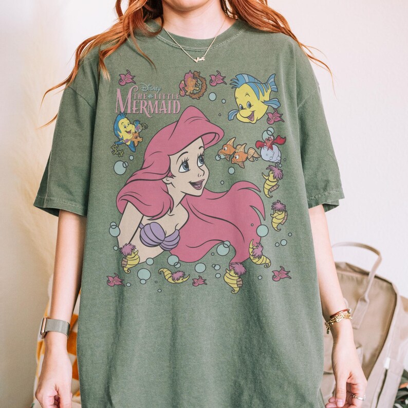 May include: Green t-shirt featuring a graphic of Ariel from Disney's The Little Mermaid. Ariel has pink hair and a purple seashell bra. The shirt also features Flounder, Sebastian, and other sea creatures. The text "Disney The Little Mermaid" is above Ariel.