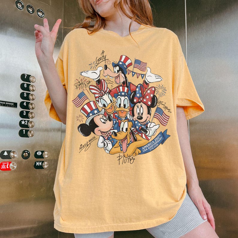 May include: Mustard yellow t-shirt with a graphic of Disney characters Mickey, Minnie, Donald, Daisy, Goofy, and Pluto in patriotic outfits. The design includes American flags and fireworks, with text "America 250th USA Anniversary".