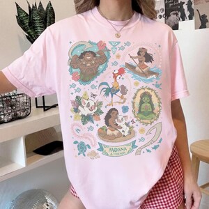 May include: A light pink t-shirt featuring colorful illustrations of characters from the animated film "Moana." The design includes images of Maui, Moana, Pua, Hei Hei, and other elements from the movie. The words "Moana & Friends" are also visible.