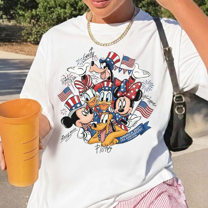 May include: White t-shirt with a vibrant graphic of Disney characters, including Mickey and Minnie Mouse, Donald and Daisy Duck, Goofy, and Pluto, celebrating the USA's 250th Anniversary. The design incorporates American flags.