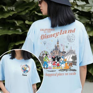 K&ouml;nnte beinhalten: Hellblaues T-Shirt mit einem Disneyland-Motiv auf der R&uuml;ckseite, einschlie&szlig;lich des Textes "CALIFORNIA Est. 1955 Disneyland happiest place on earth". Auf der Vorderseite befindet sich eine Donald Duck-Grafik und der Text "NAME".