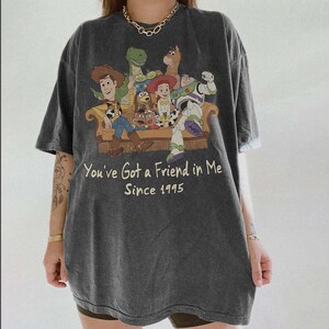 May include: A charcoal gray short-sleeve t-shirt featuring a graphic print of Toy Story characters sitting on a couch. The text reads, You've Got a Friend in Me Since 1995. The shirt has a crew neckline and a relaxed, loose-fitting silhouette.