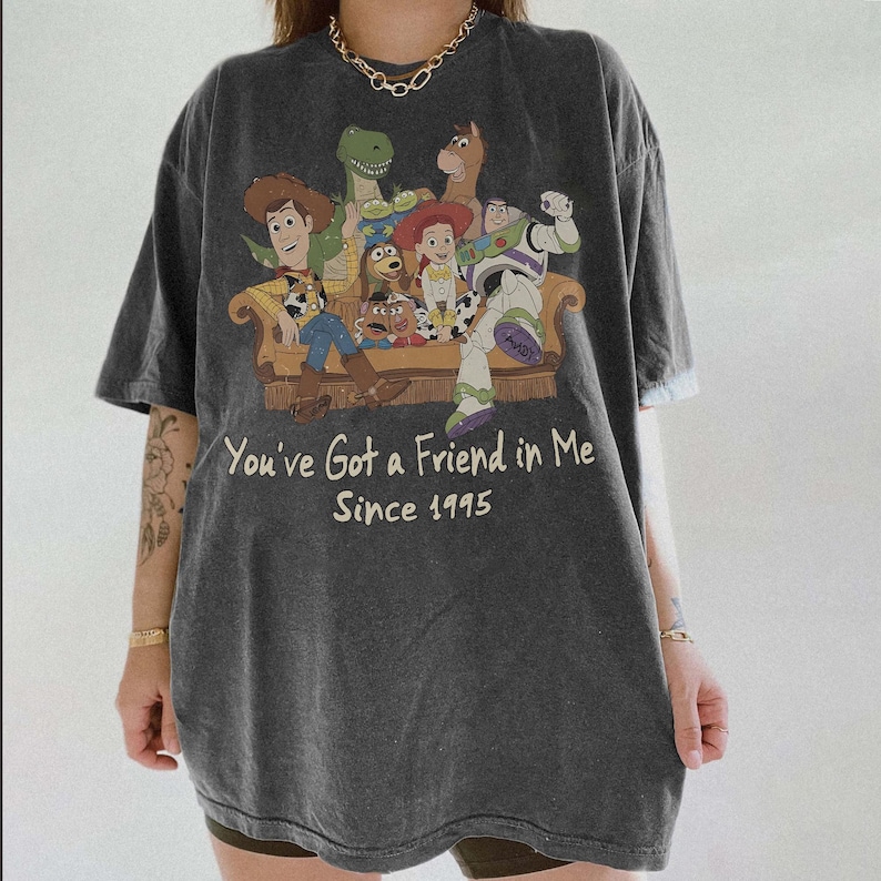 May include: A charcoal gray short-sleeve t-shirt featuring a graphic print of Toy Story characters sitting on a couch. The text reads, You've Got a Friend in Me Since 1995. The shirt has a crew neckline and a relaxed, loose-fitting silhouette.