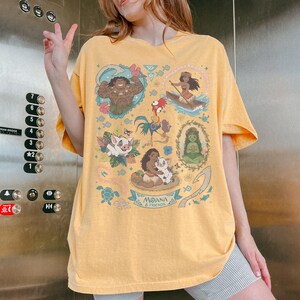 May include: Yellow t-shirt featuring a graphic design with characters from the animated film "Moana." The design includes illustrations of Maui, Moana, Hei Hei, Pua, and other characters. The text "Moana & Friends" is also included.