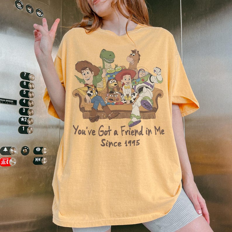 May include: A yellow short-sleeve t-shirt featuring a graphic print of Toy Story characters sitting on a couch. The text below the characters reads, "You've Got a Friend in Me Since 1995." The shirt has a crew neckline and a relaxed, loose fit.