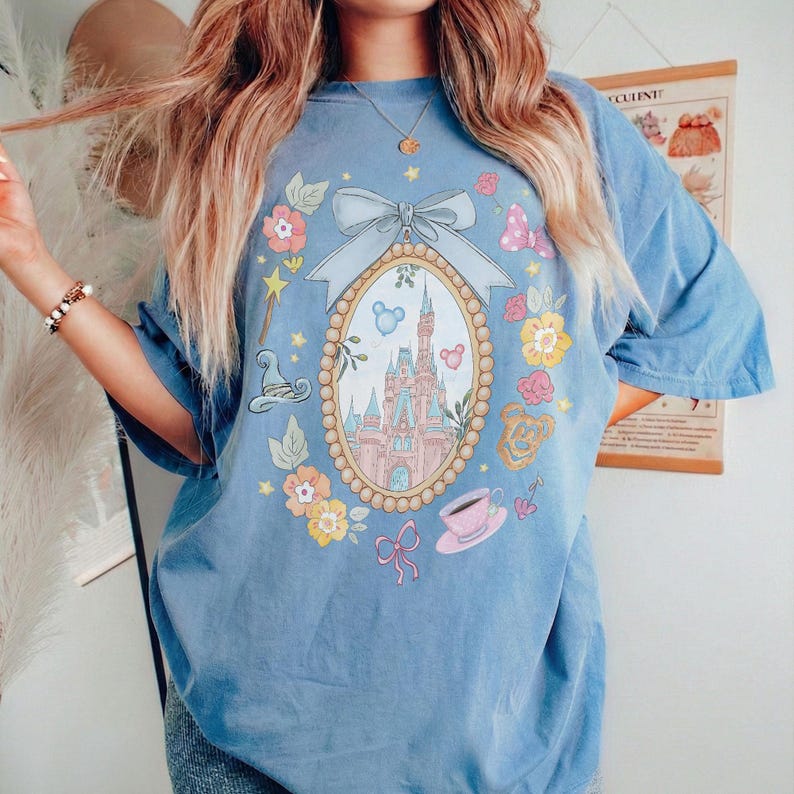 May include: A light blue t-shirt with a central oval design depicting a castle, surrounded by flowers and whimsical motifs. The shirt has short sleeves and a relaxed fit, suitable for everyday wear.