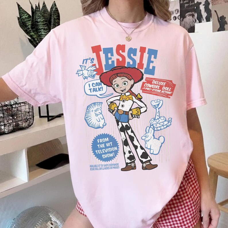 May include: A light pink t-shirt featuring a graphic of the character Jessie from the animated film Toy Story. The design includes the text "JESSIE", "I CAN TALK!", and "DELUXE COWGIRL DOLL". Additional graphics include a hedgehog, snake, and a rabbit.