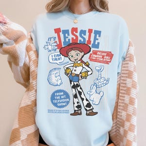 May include: Light blue t-shirt featuring a graphic of Jessie from the Toy Story franchise. The design includes the text "JESSIE", "I CAN TALK!", and "DELUXE COWGIRL DOLL". Additional graphics of a snake, a rabbit, and a hedgehog are also present.